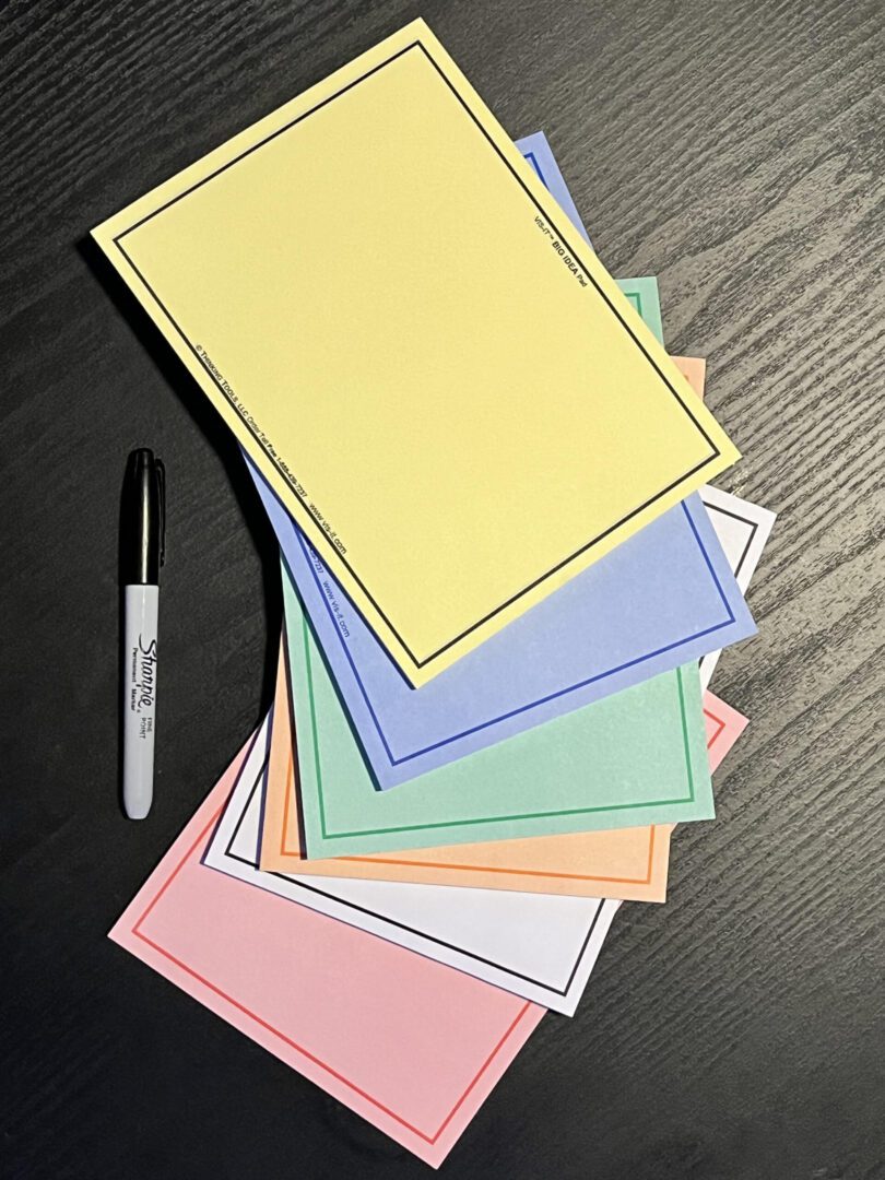 A stack of SIX-COLOR Set of VIS-IT™ 8x6 in. Big Idea Pads (300 sheets total) and a marker on a table.